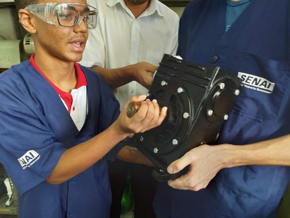Amós aprendiz no SENAI, de uniforme, segurando uma peça usinada