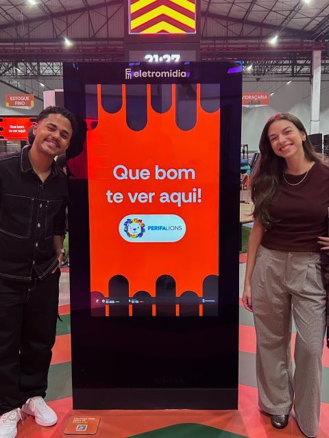 Amós e Lorena ao lado de um totem da Eletromidia com a mensagem Que bom te ver aqui, PerifaLions