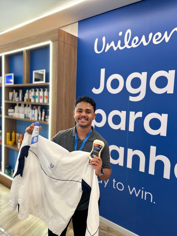 Amós segurando a jaqueta da Unilever, um Dove e uma Hellmann’s em frente ao painel Joga para ganhar