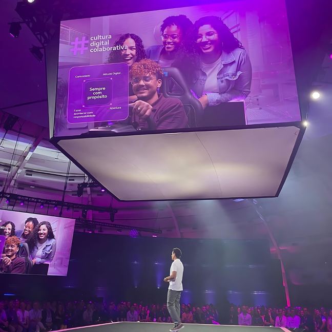 Amós no palco de um evento interno da Vivo, com telão roxo